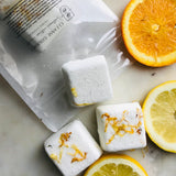 Five white cubes of Invigorate Shower Steamers Organic Citrus by Cleanse Gourmet with orange slices on natural counter. 