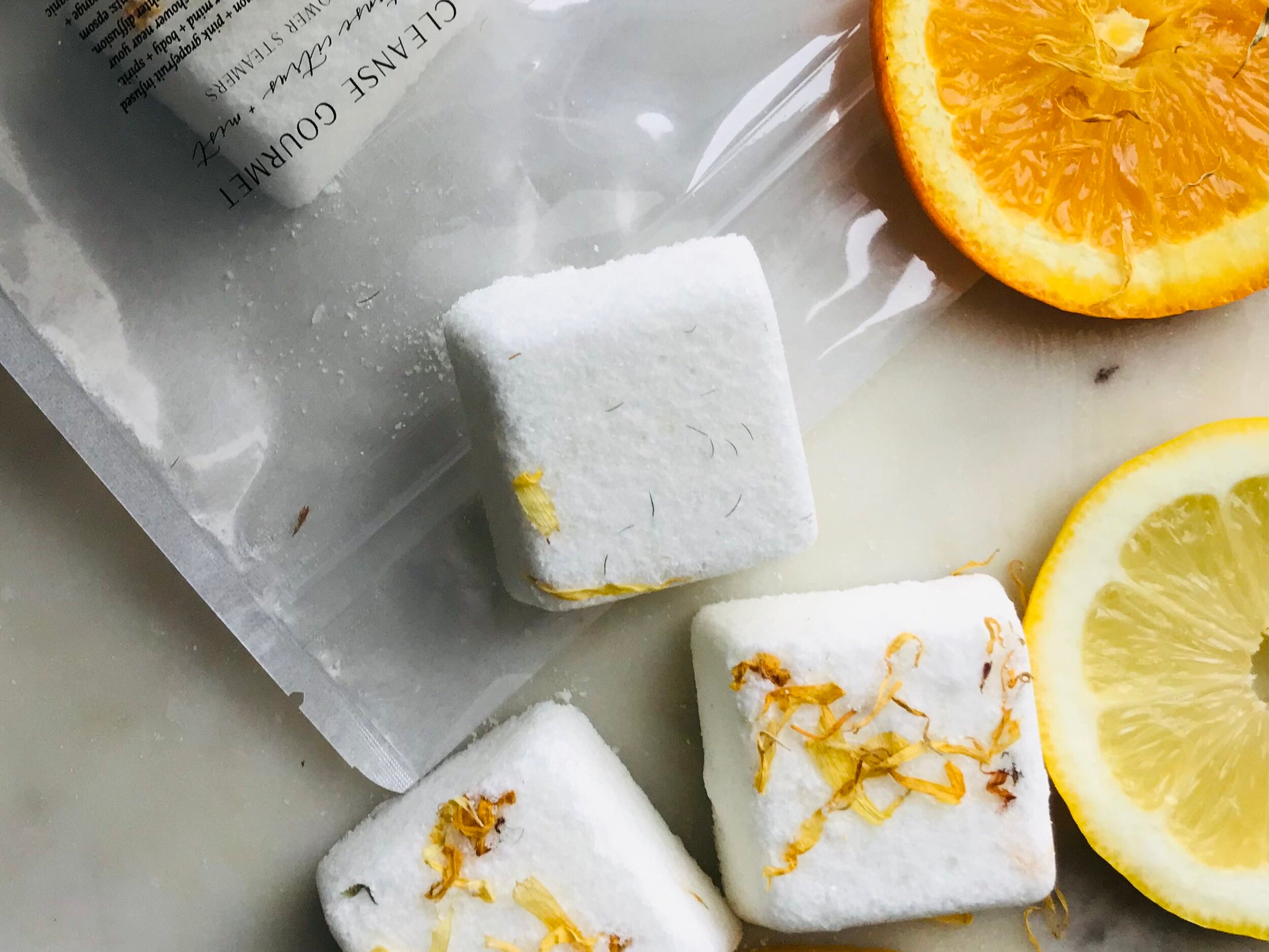 Five white cubes of Invigorate Shower Steamers Organic Citrus by Cleanse Gourmet with orange slices on natural counter. 