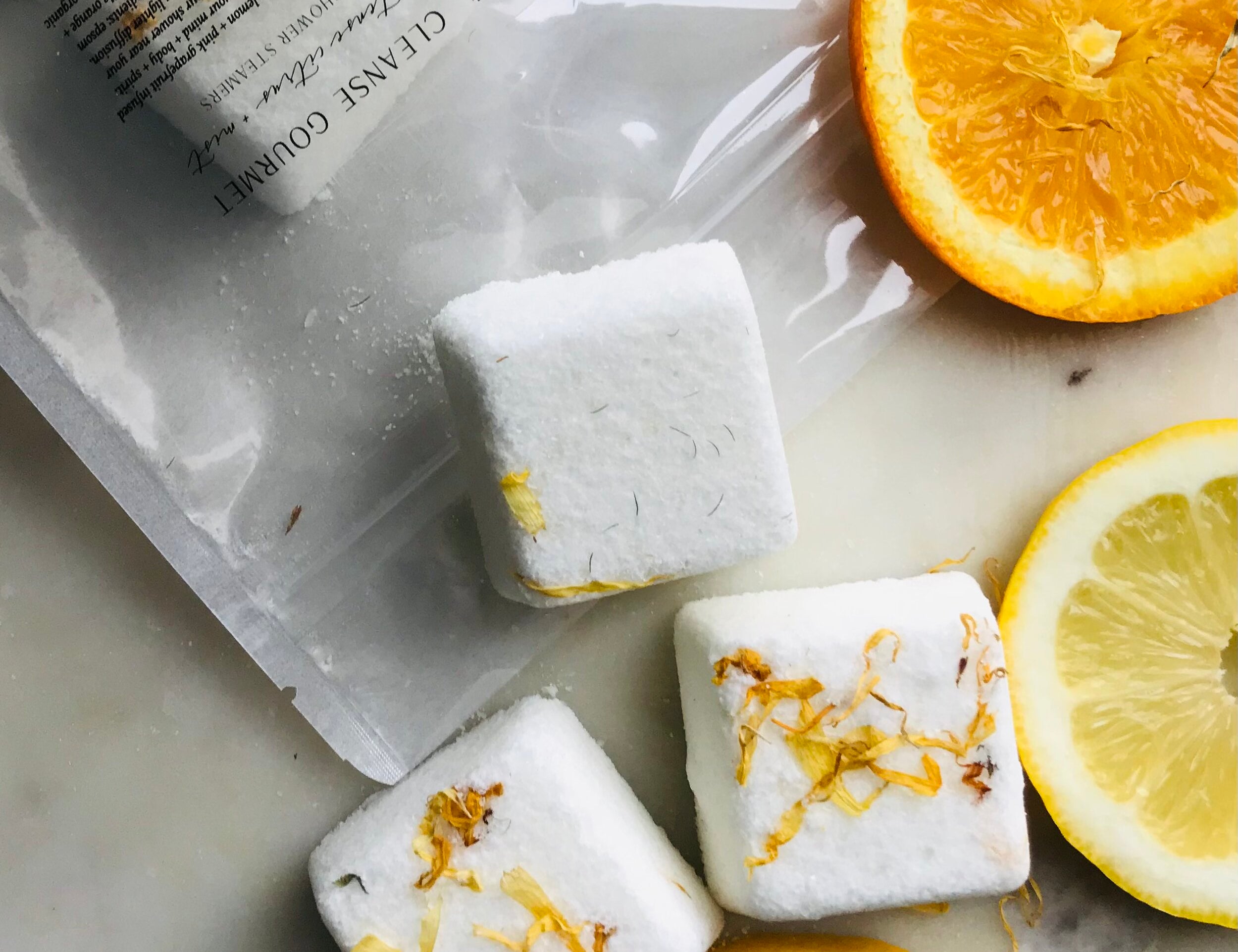 Five white cubes of Invigorate Shower Steamers Organic Citrus by Cleanse Gourmet with orange slices on natural counter. 