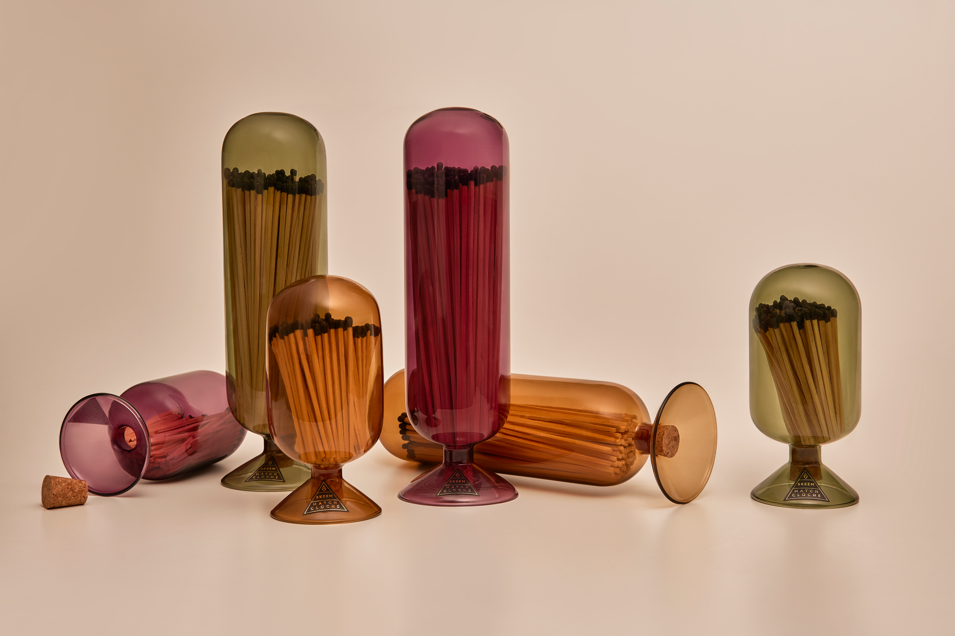 Match Cloches by Skeem in jewel-toned vintage glass domes fitted with cork. Comes in olive green, amber sun, and mulberry wine and holds 120 matches. Six in tall and short sizes over tan background.