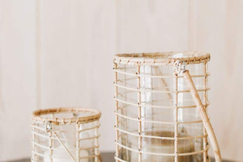 Two Glass Lieto Lanterns with rattan exterior and handle in natural light wood tone holding white candles and set in airy home