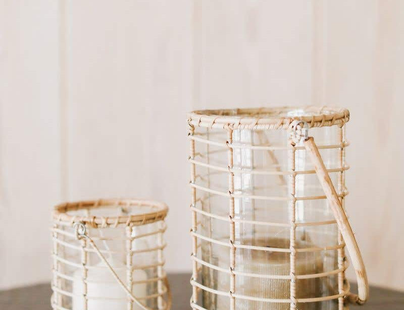 Two Glass Lieto Lanterns with rattan exterior and handle in natural light wood tone holding white candles and set in airy home