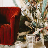 Two Glass Lieto Lanterns with rattan exterior and handle in natural light wood tone holding white candles and set by red velvet couch and dried bouquet