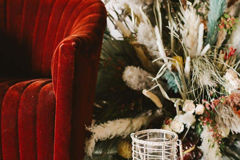 Two Glass Lieto Lanterns with rattan exterior and handle in natural light wood tone holding white candles and set by red velvet couch and dried bouquet