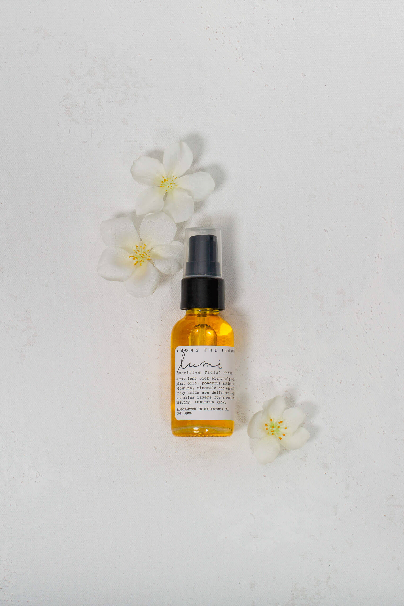 Golden Lumi Facial Serum in clear bottle with black cap surrounded by white flowers