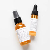 Two amber glass dropper and spray bottles with white labels, photographed on a clean white background. The label text appears to read 'Among the Flowers' and 'lumi'.
