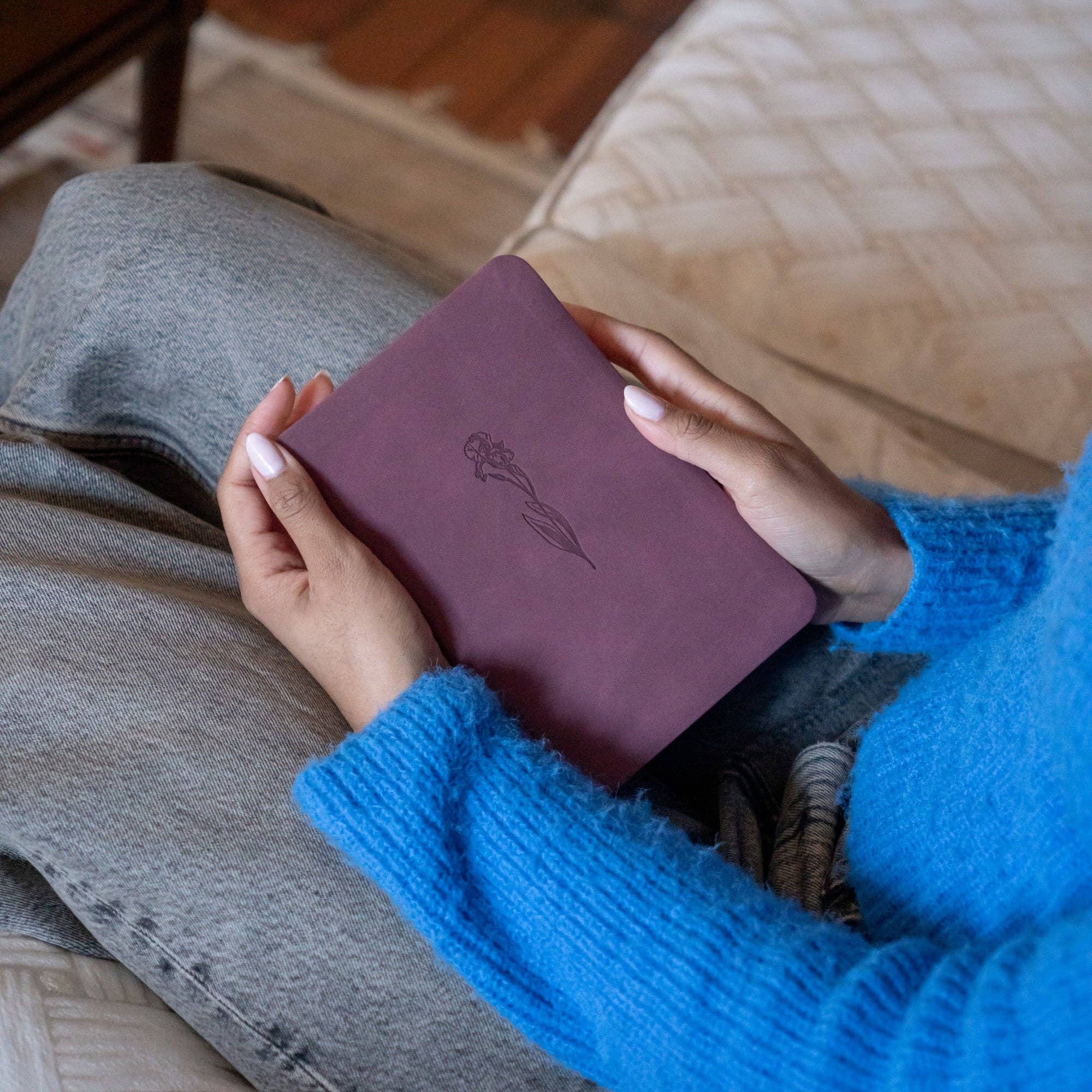 Person holding a maroon leather notebook with a logo on a wooden floor.