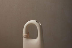 Palau Tan Vase matte finish with natural bisque cermaic, unique handle feature and wrapped rattan coil.  Sitting on stone table and backed by brown wall.