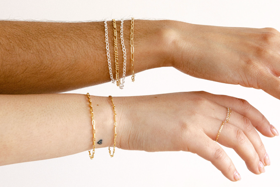 Two human arms and hands wearing delicate gold jewelry against a plain light background, including stacked chain bracelets on the upper wrist and a few bracelets and a ring on the lower wrist and hand.