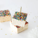 Three squares of Rainbow Sprinkles Crispy Cake by Lolli & Pops and a golden candle and golden star confetti. 