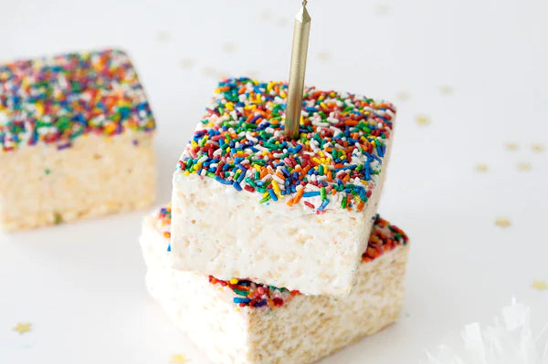 Three squares of Rainbow Sprinkles Crispy Cake by Lolli & Pops and a golden candle and golden star confetti. 