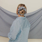 Person wearing a striped shirt with a blue scrunchie on their head, standing in front of a striped fabric backdrop.
