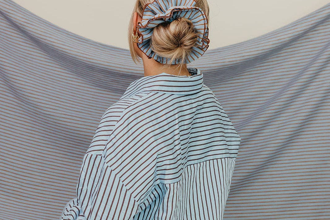Person wearing a striped shirt with a blue scrunchie on their head, standing in front of a striped fabric backdrop.