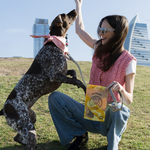 Woman playing with a dog in an open field with buildings in the background