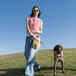 Woman standing on a grassy hill with a dog, holding a book, against a clear blue sky.