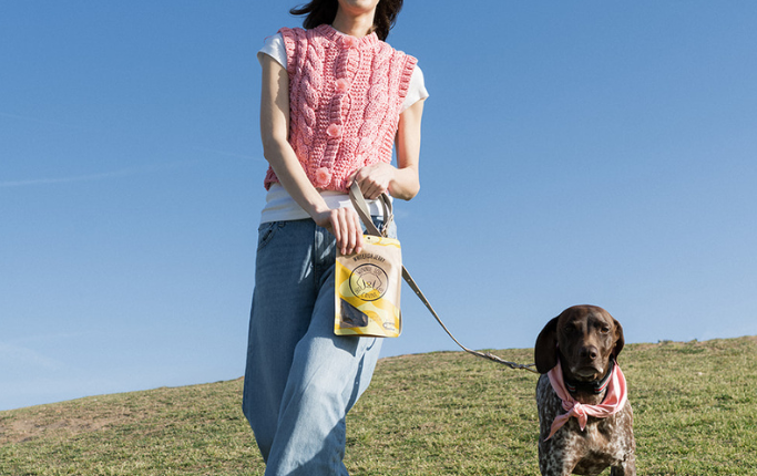 Woman standing on a grassy hill with a dog, holding a book, against a clear blue sky.
