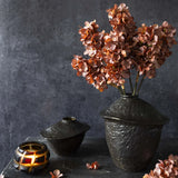 Abigail Ahern Artificial Cone Hydrangea in Antique Pink, three stems in a black ceramic vase on a dark table with a candle