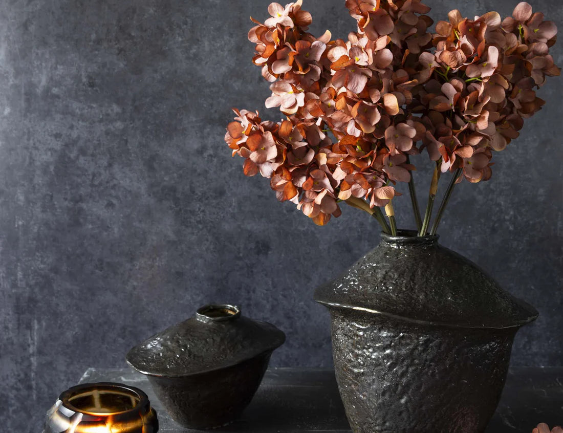 Abigail Ahern Artificial Cone Hydrangea in Antique Pink, three stems in a black ceramic vase on a dark table with a candle