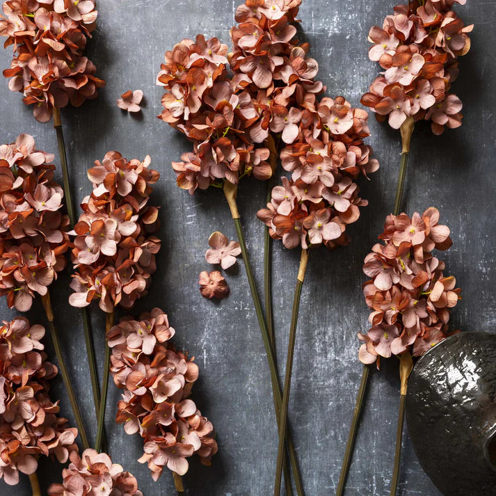 Abigail Ahern Artificial Cone Hydrangea in Antique Pink, several rust-pink faux stems laid on a dark grey surface