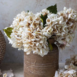 Abigail Ahern Artificial Hydrangea in Morning Mist, full cream blooms in a brown ceramic vase beside woven pots