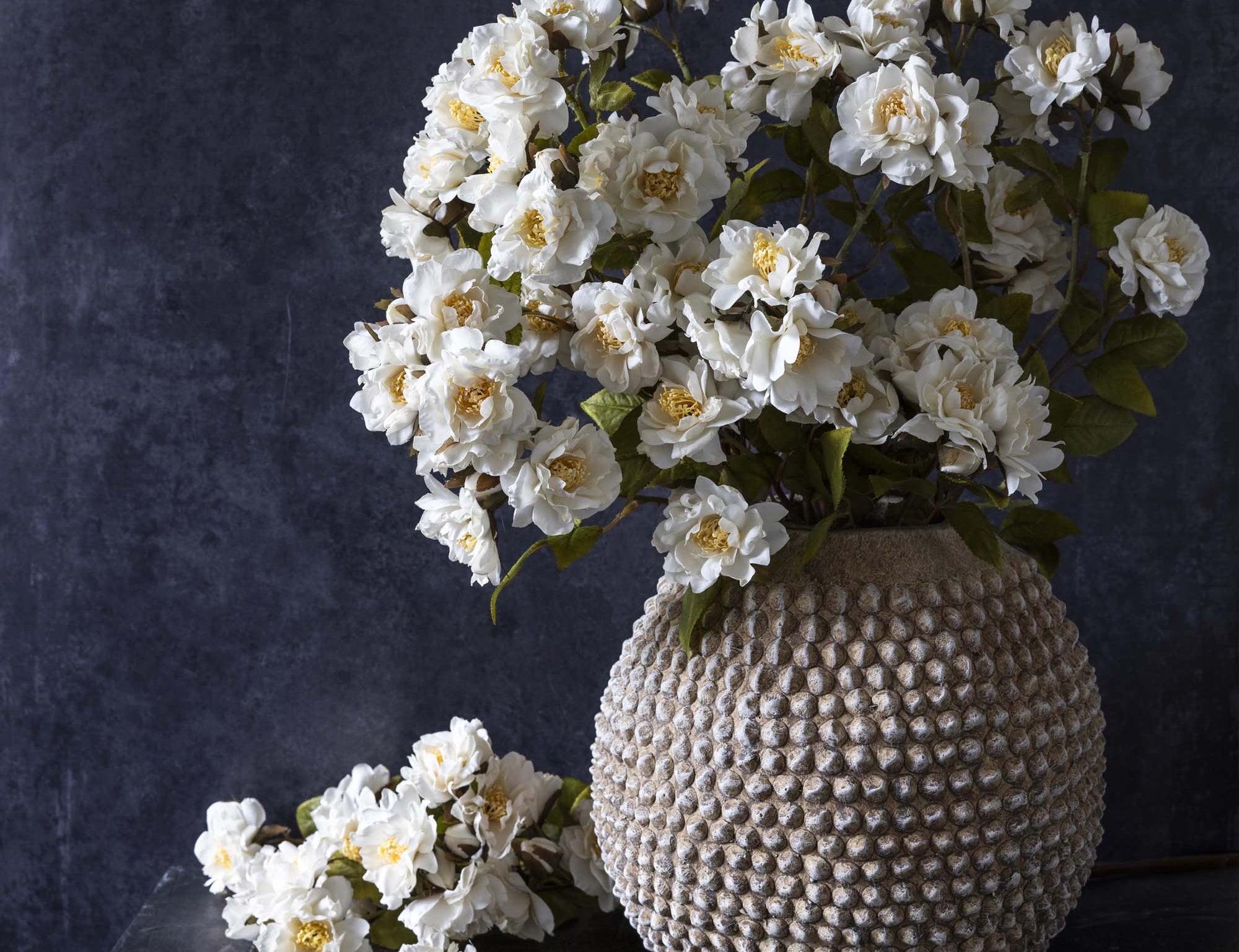 Abigail Ahern Artificial Rose Cream Spray, full bouquet of white faux roses in a textured stone vase on a dark background
