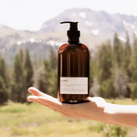 Hand holding the Among the Flowers Botanical Aloe 8oz Lotion in Driftwood, an amber pump bottle, with mountains behind