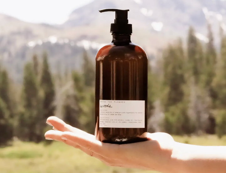 Hand holding the Among the Flowers Botanical Aloe 8oz Lotion in Driftwood, an amber pump bottle, with mountains behind
