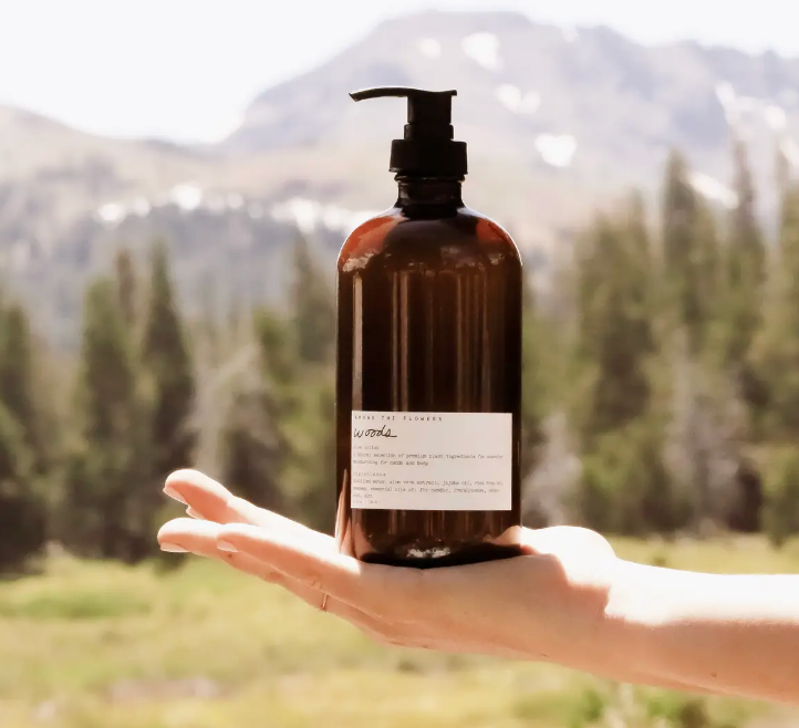 Hand holding the Among the Flowers Botanical Aloe 8oz Lotion in Driftwood, an amber pump bottle, with mountains behind