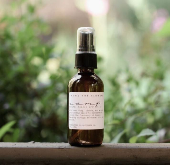Among the Flowers Camp Mist Insect Shield amber spray bottle on a ledge with greenery behind
