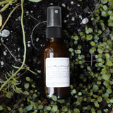 Among the Flowers Camp Mist Insect Shield amber spray bottle resting on a bed of green ground cover