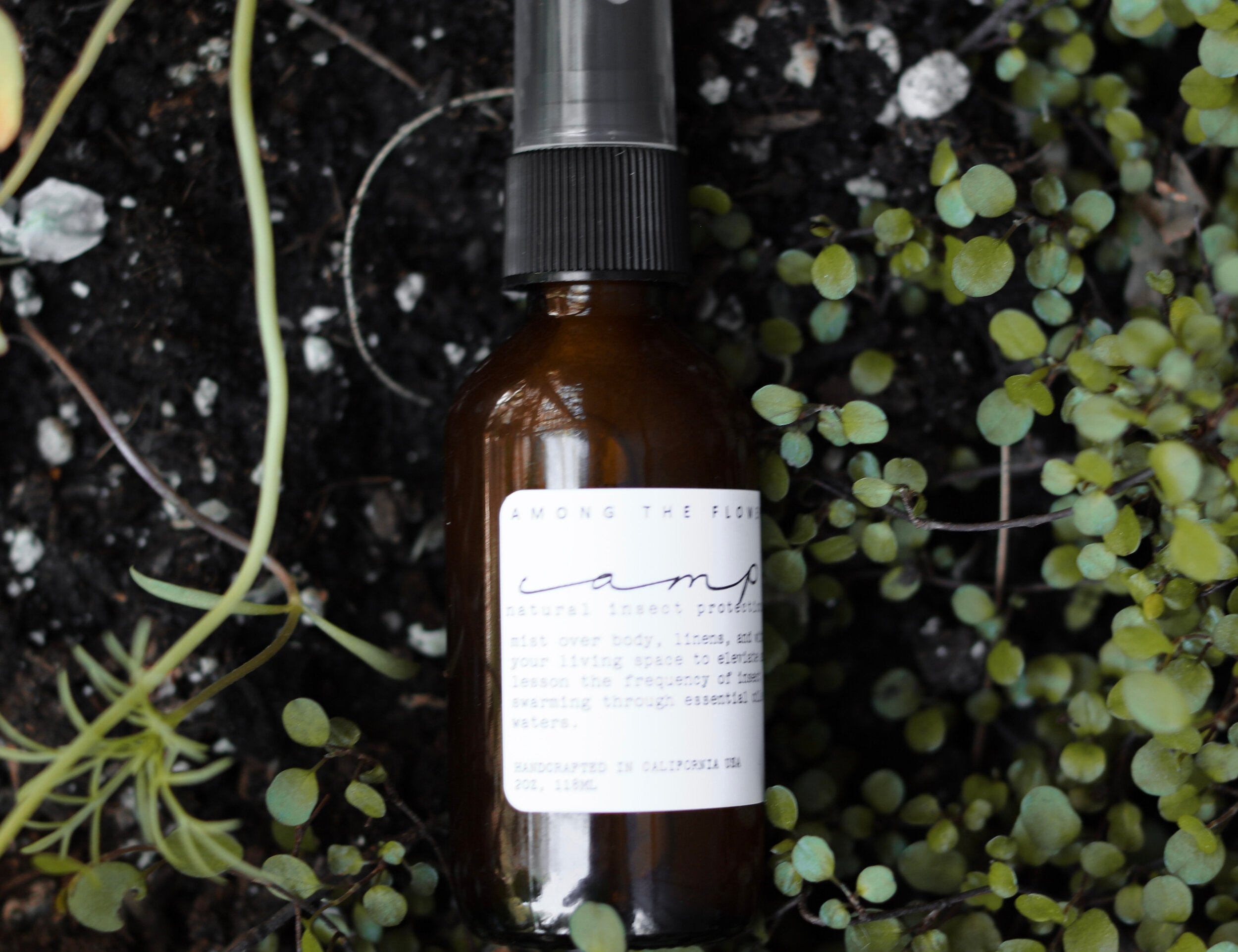 Among the Flowers Camp Mist Insect Shield amber spray bottle resting on a bed of green ground cover