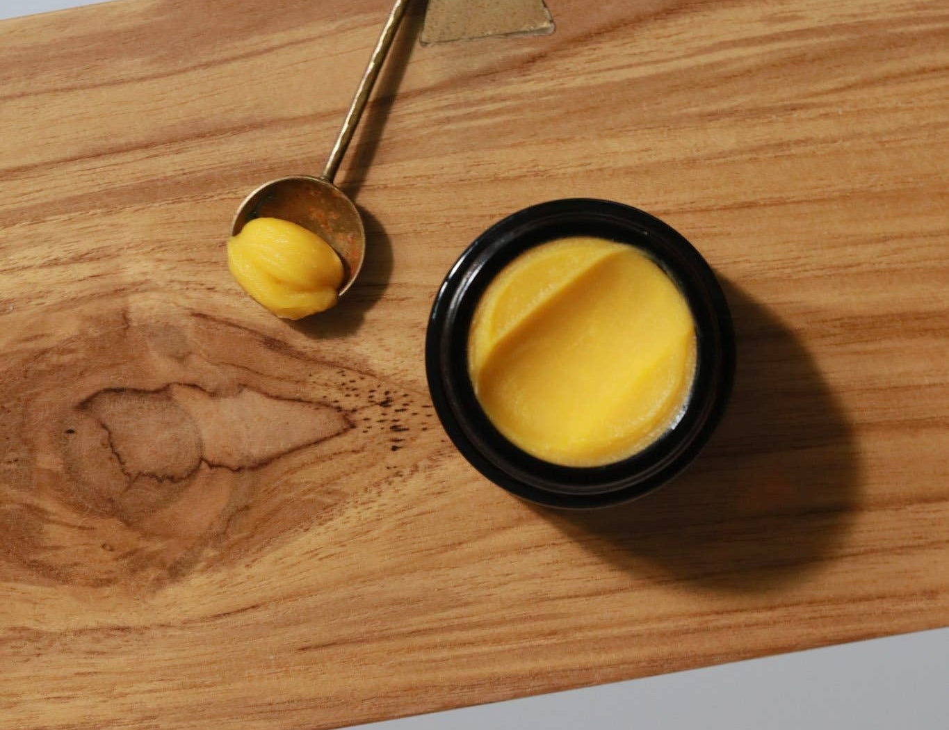 Among the Flowers Gold Balancing Face Balm in a black jar on a wooden board with a gold spoon of balm