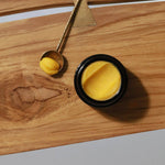 Among the Flowers Gold Balancing Face Balm in a black jar on a wooden board with a gold spoon of balm