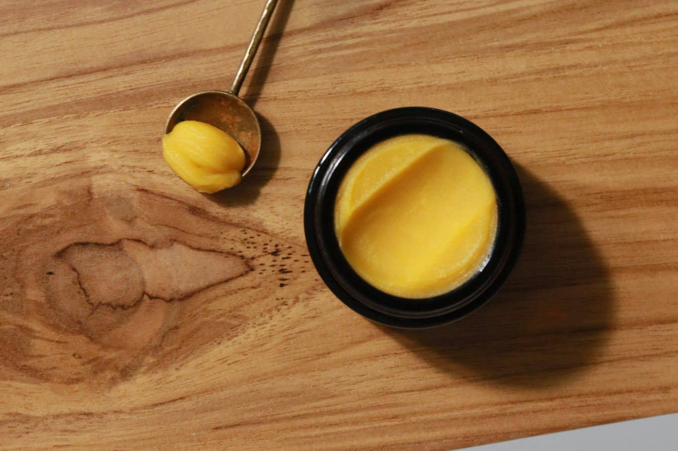 Among the Flowers Gold Balancing Face Balm in a black jar on a wooden board with a gold spoon of balm