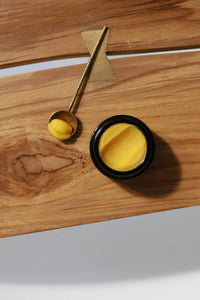 Among the Flowers Gold Balancing Face Balm in a black jar on a wooden board with a gold spoon of balm