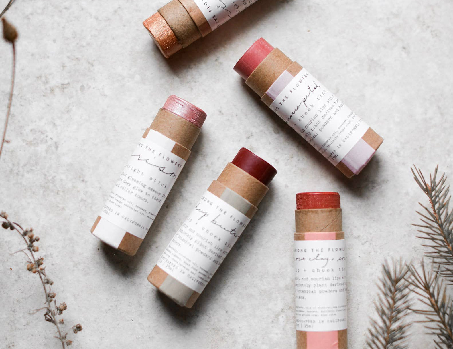 Among the Flowers Lip and Cheek Tint tubes scattered on a stone surface with dried botanicals