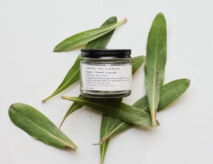 Among the Flowers Purify Face Mask in a small glass jar with a black lid, surrounded by green leaves