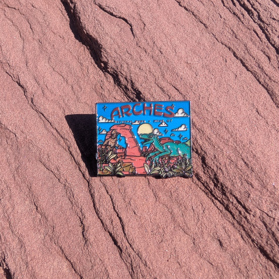 Square Arches enamel pin with Bigfoot and a green raptor beside Delicate Arch under a blue sky, lying on red sandstone in Moab, Utah.