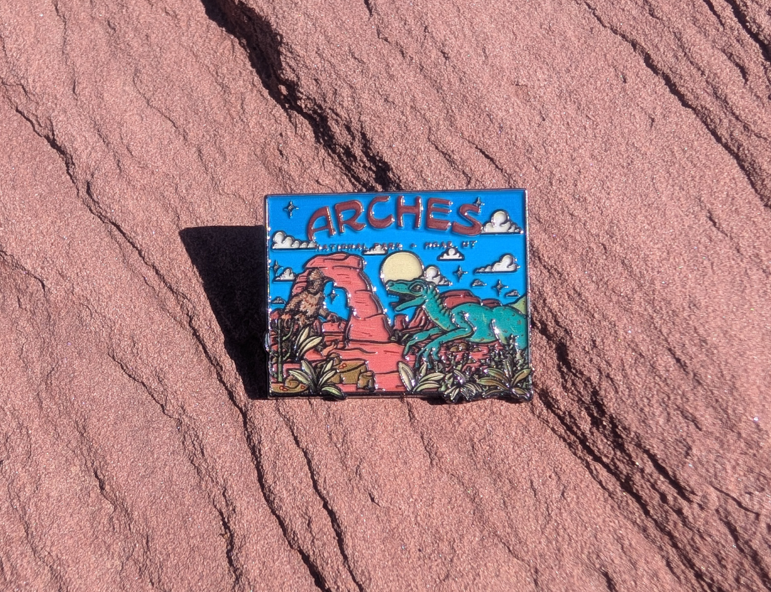 Square Arches enamel pin with Bigfoot and a green raptor beside Delicate Arch under a blue sky, lying on red sandstone in Moab, Utah.