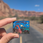 Hand holding the Arches Bigfoot and raptor enamel pin on a road through the Moab red rock cliffs.