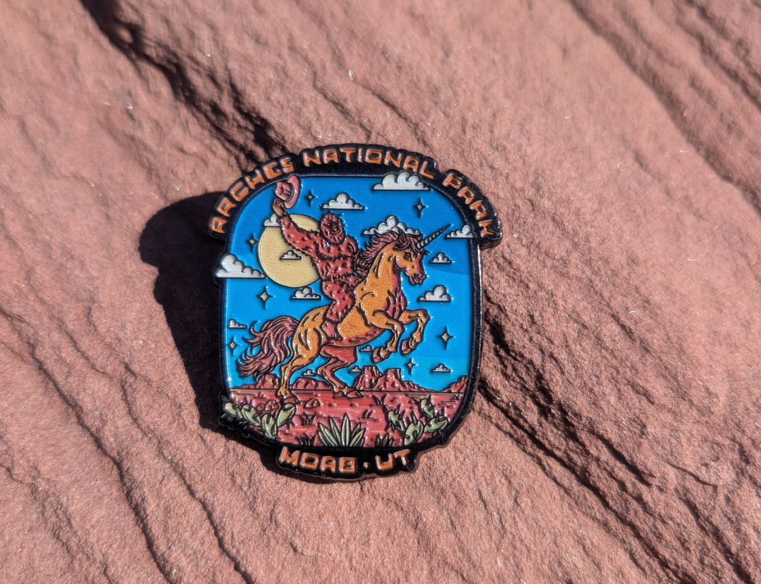 Arches National Park Moab UT enamel pin with Bigfoot riding a rearing unicorn through red rock, lying on red sandstone in Moab, Utah.