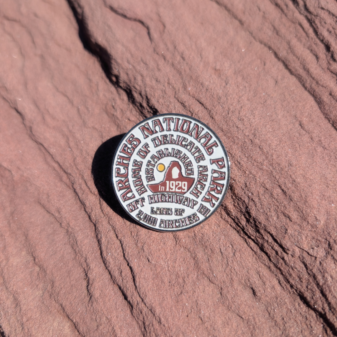 Round white and maroon Arches National Park enamel pin lettered with 1929 and Delicate Arch, lying on red sandstone in Moab, Utah.
