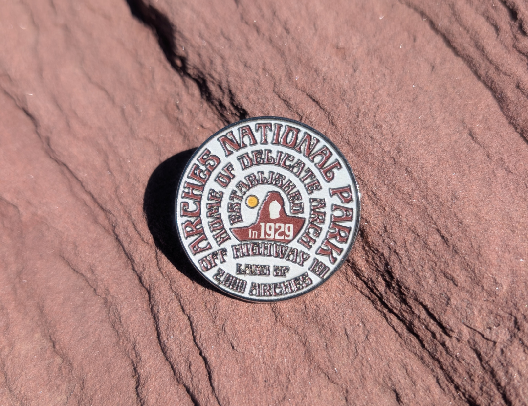 Round white and maroon Arches National Park enamel pin lettered with 1929 and Delicate Arch, lying on red sandstone in Moab, Utah.