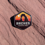 Hexagon Arches National Park enamel pin with Delicate Arch silhouetted against a yellow and orange sunset, lying on red sandstone in Moab, Utah.