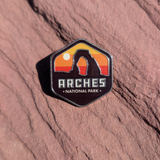 Hexagon Arches National Park enamel pin with Delicate Arch silhouetted against a yellow and orange sunset, lying on red sandstone in Moab, Utah.