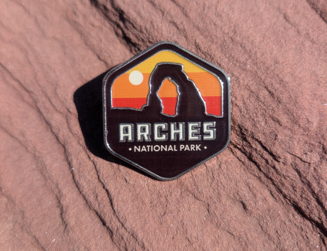 Hexagon Arches National Park enamel pin with Delicate Arch silhouetted against a yellow and orange sunset, lying on red sandstone in Moab, Utah.