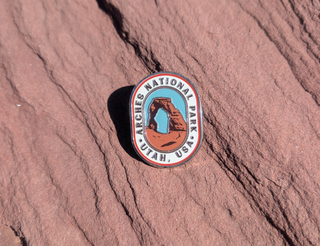 Oval Arches National Park Utah USA enamel pin with Delicate Arch against a blue sky, lying on red sandstone in Moab, Utah.