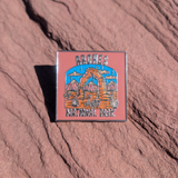 Square Arches National Park enamel pin with Delicate Arch line art on a dusty pink sky, lying on red sandstone in Moab, Utah.