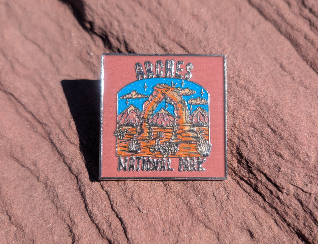 Square Arches National Park enamel pin with Delicate Arch line art on a dusty pink sky, lying on red sandstone in Moab, Utah.