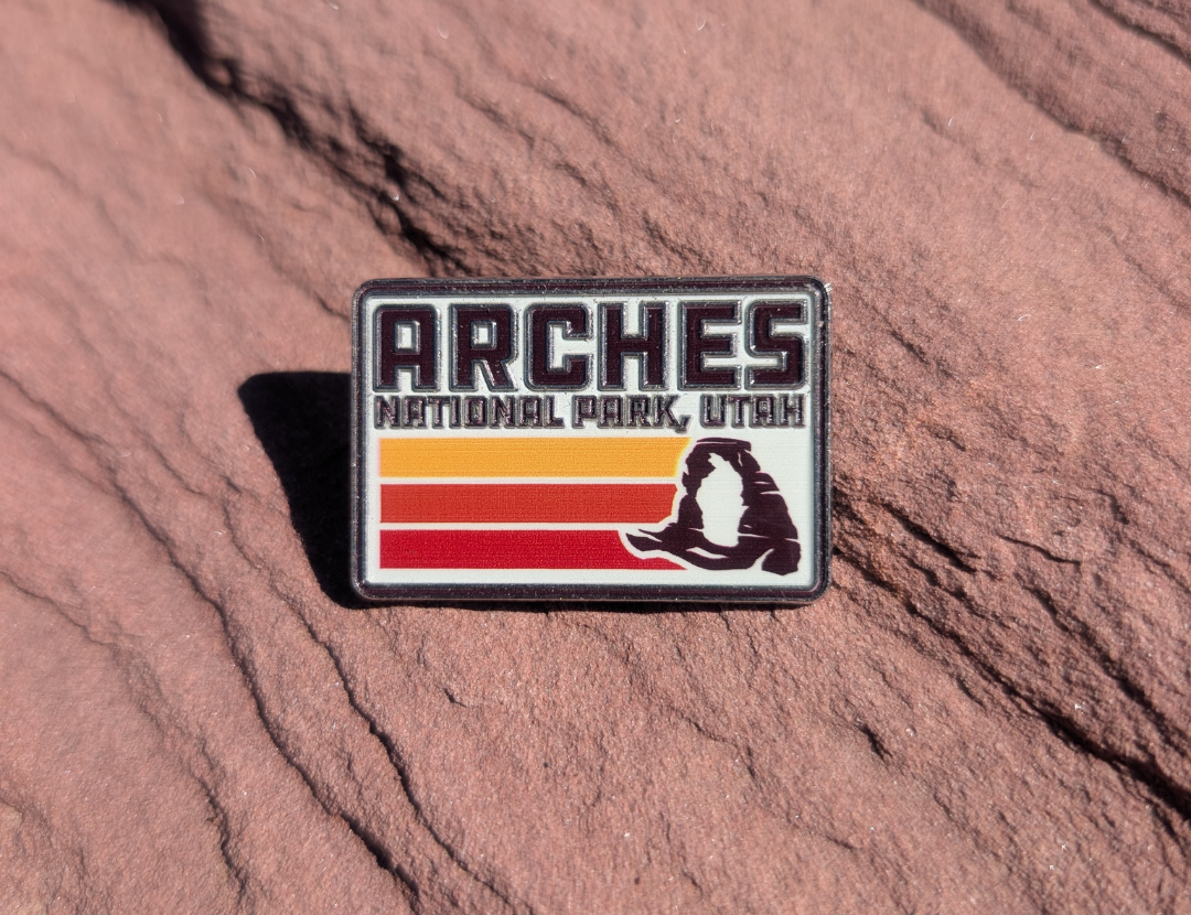Rectangular Arches National Park Utah enamel pin with retro orange and red stripes and Delicate Arch, lying on red sandstone in Moab, Utah.
